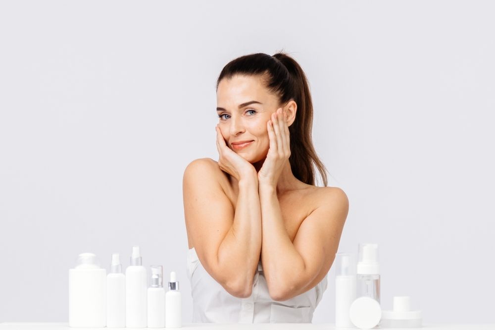 Woman with radiant skin smiles, touches cheeks, surrounded by white skincare products.
