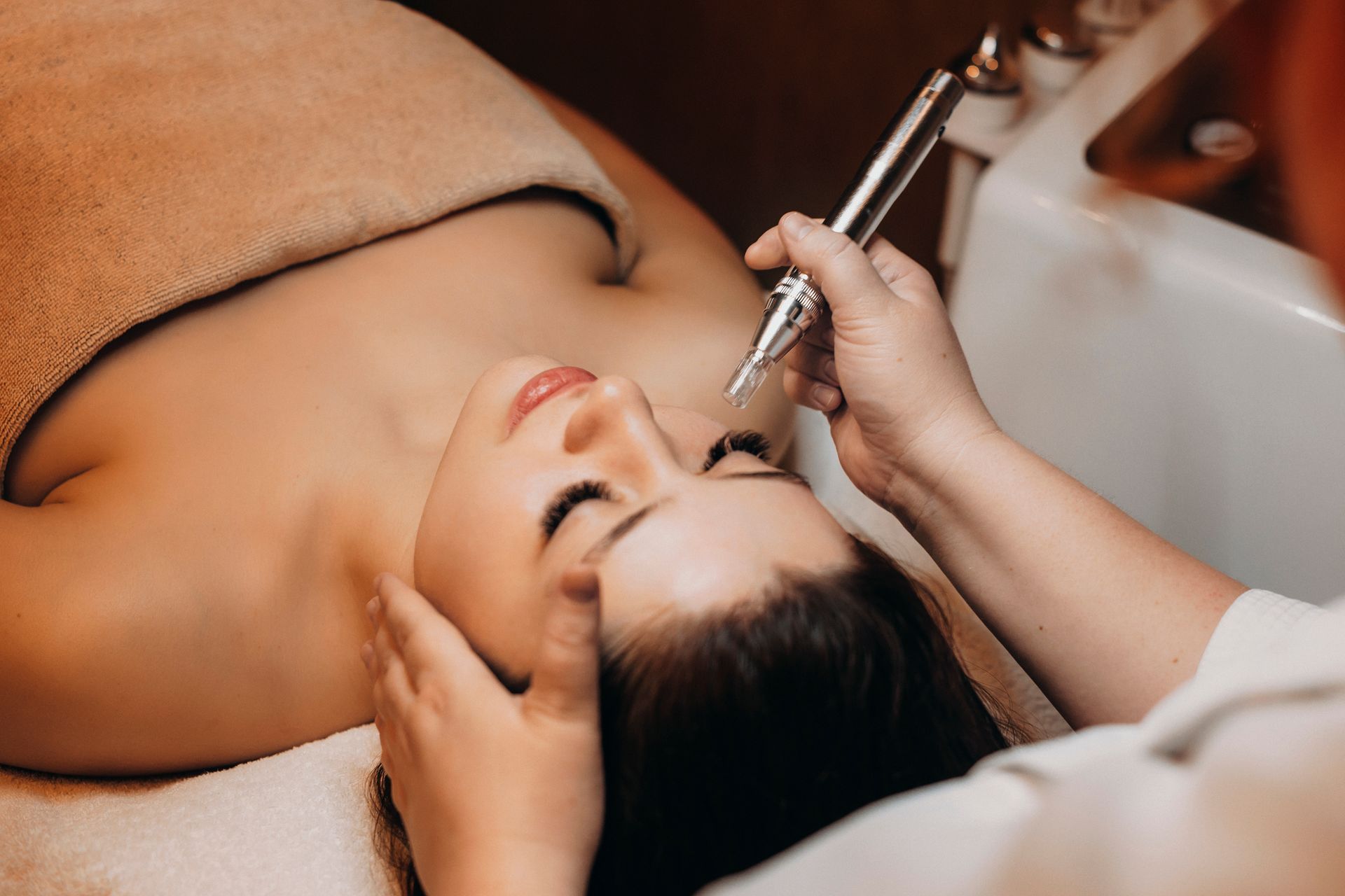 Woman receiving facial treatment with a metal tool, eyes closed, in a spa setting.