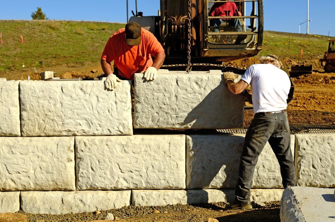 Two workers placing large, light-colored stone blocks to build a wall on a construction site.