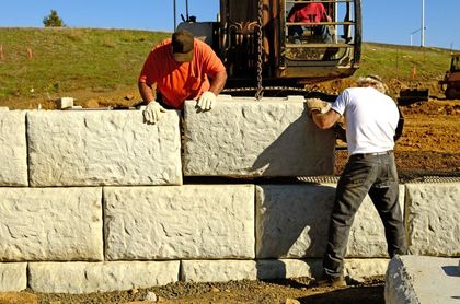 Two workers placing large, light-colored stone blocks to build a wall on a construction site.