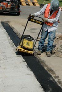 Construction worker compacting asphalt with a vibratory plate compactor in a road.