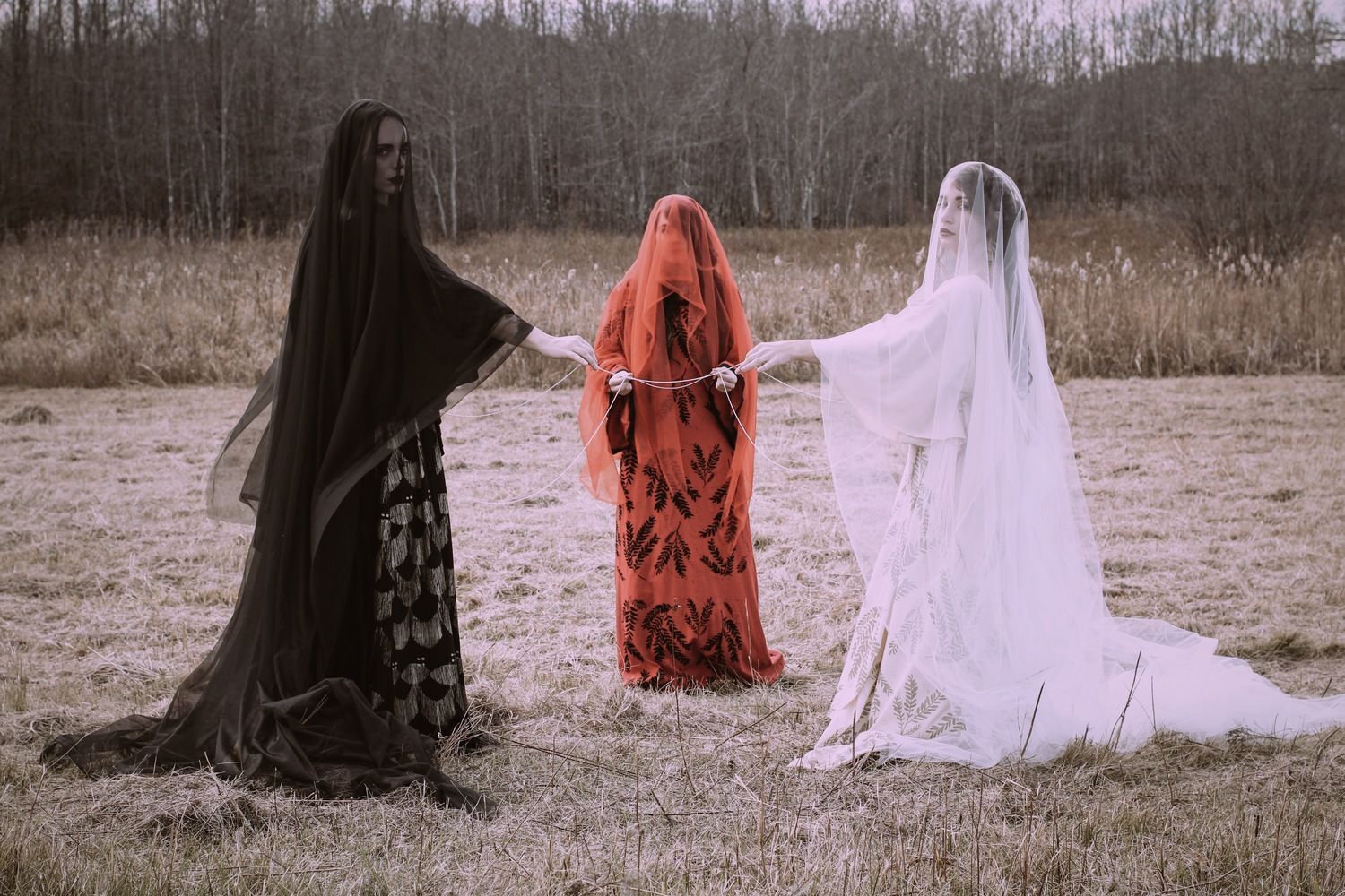 Three women in long dresses for ceremony are standing in a field holding hands.