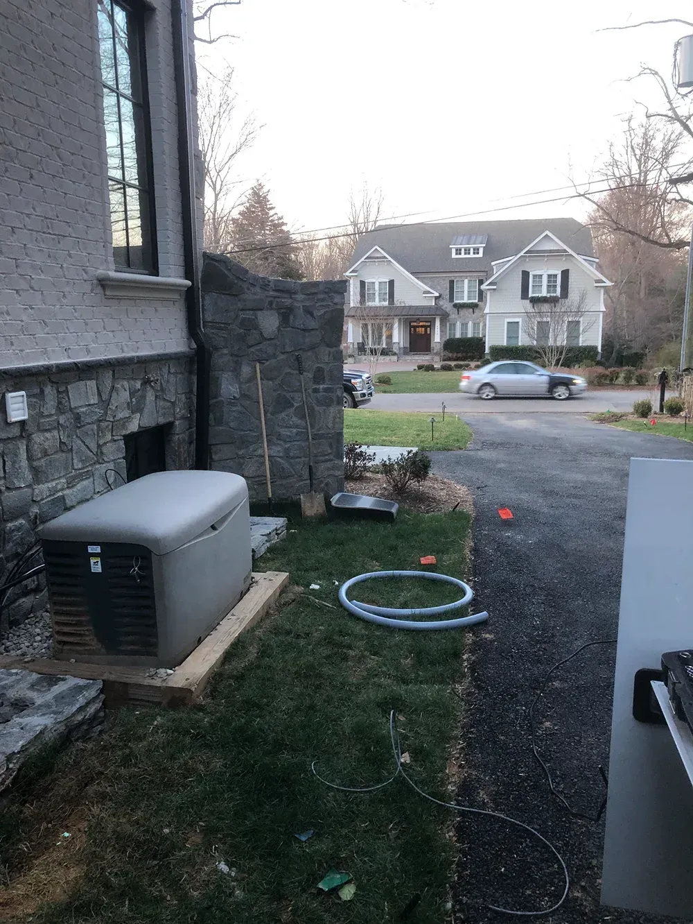 Side yard with HVAC unit, hose on gravel, and a residential street with houses and parked car beyond