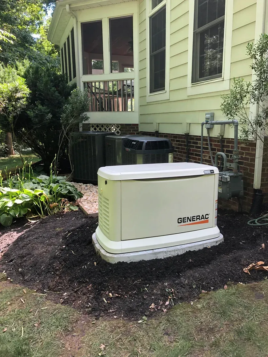 Beige generator on a mulch bed beside a green house and shrubs.