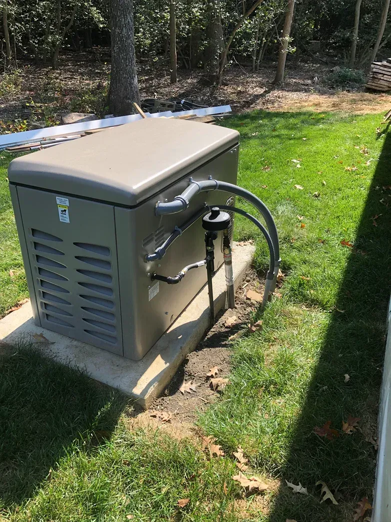 Outdoor generator unit beside a house, with hoses and cables on a small concrete pad in a grassy yard