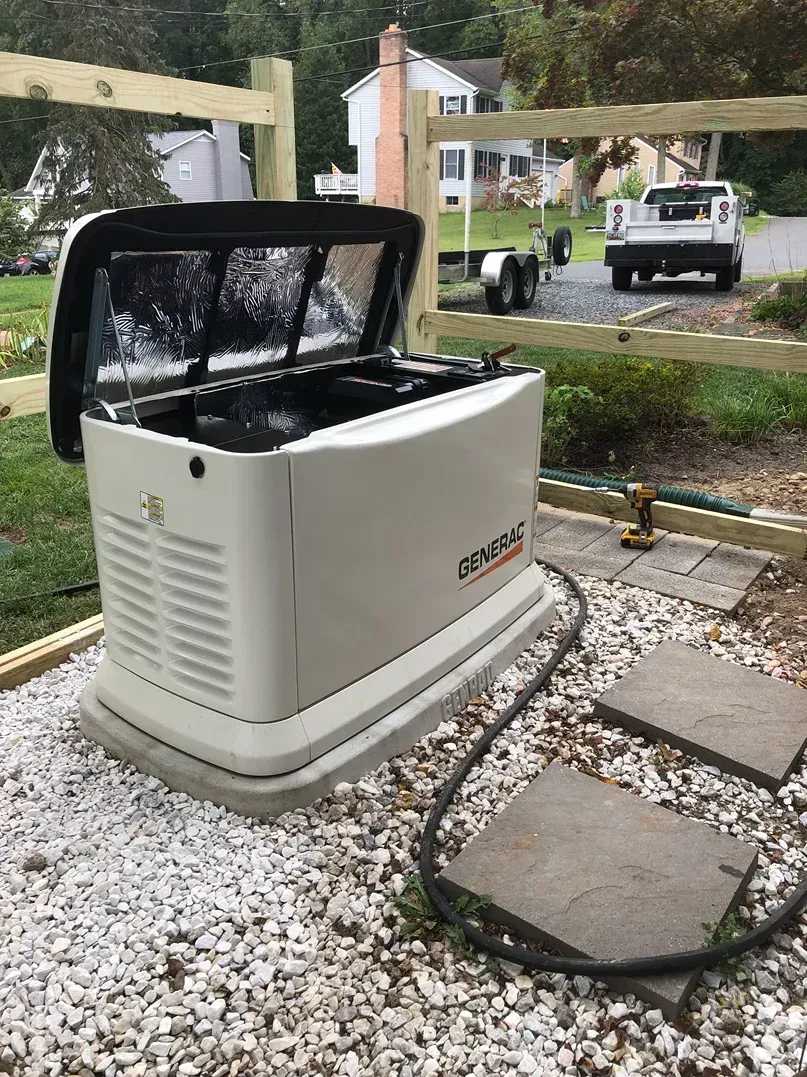 Outdoor standby generator with open lid on gravel beside a driveway and yard, with a truck in the background