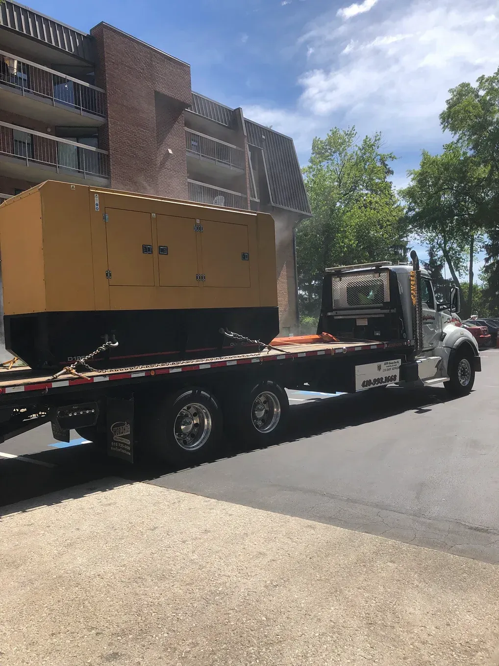 Flatbed tow truck hauling large yellow equipment on a street beside a brick building.