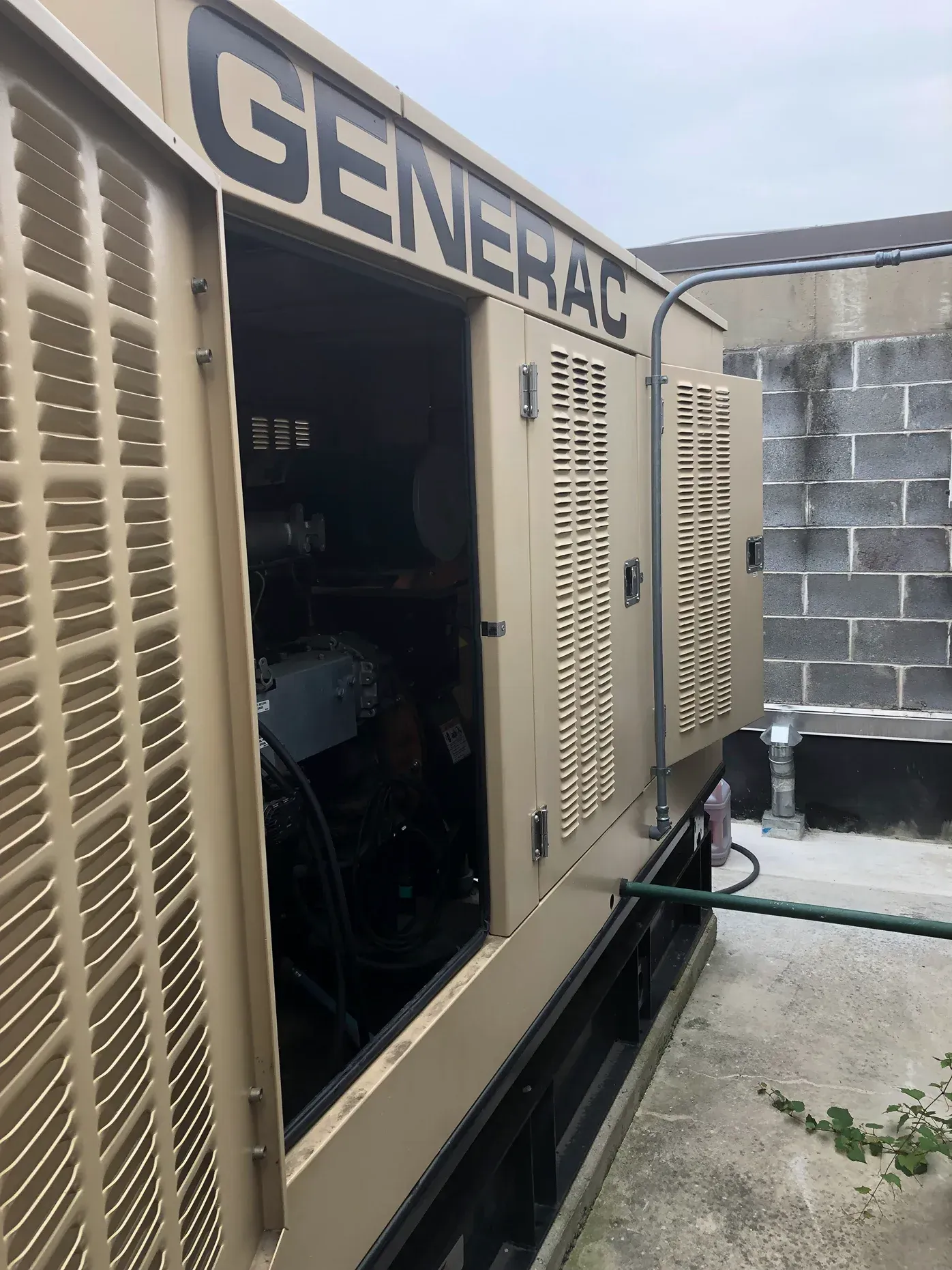 Open Generac generator unit with beige vents beside a building wall and concrete walkway.