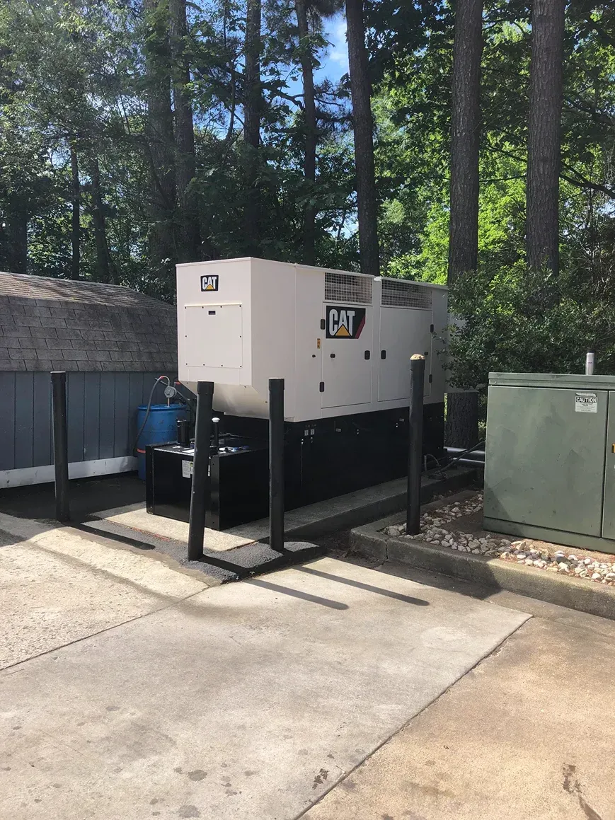 Industrial generator beside a building, fenced off with black bollards and surrounded by trees.