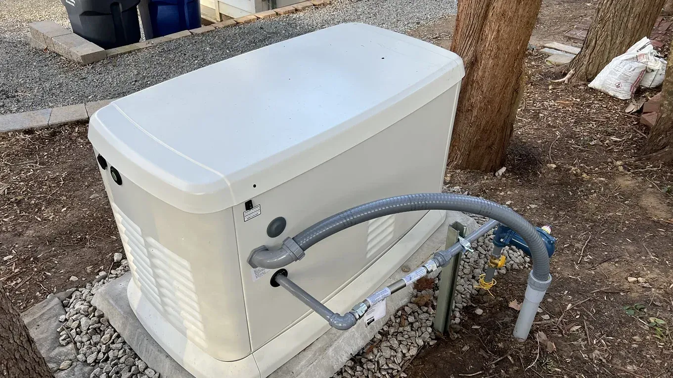 Outdoor white utility box with attached gray conduit and pipes beside a tree on gravel.
