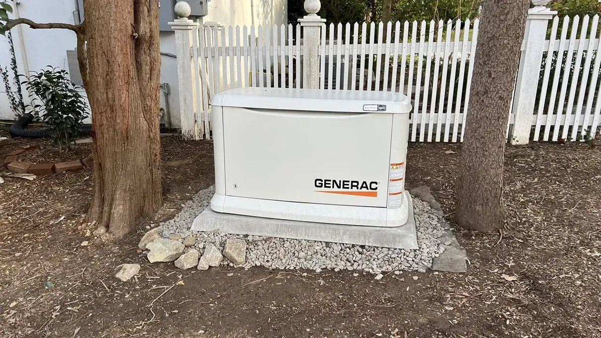 White Generac generator on gravel beside a tree and white picket fence in a yard