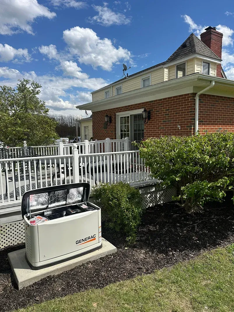 White generator on concrete pad beside a white fence and brick house under a blue sky
