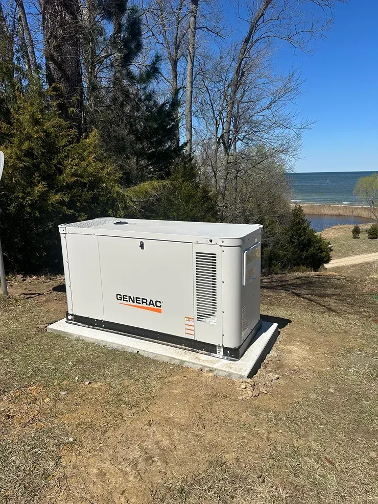 White Generac generator on a concrete pad in a grassy yard near trees and a lake.