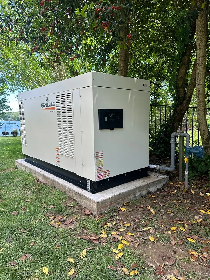 White industrial generator on a concrete pad beside trees and a utility line in a grassy area