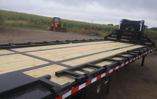 A trailer with a wooden deck is parked in a dirt field with a tractor in the background.