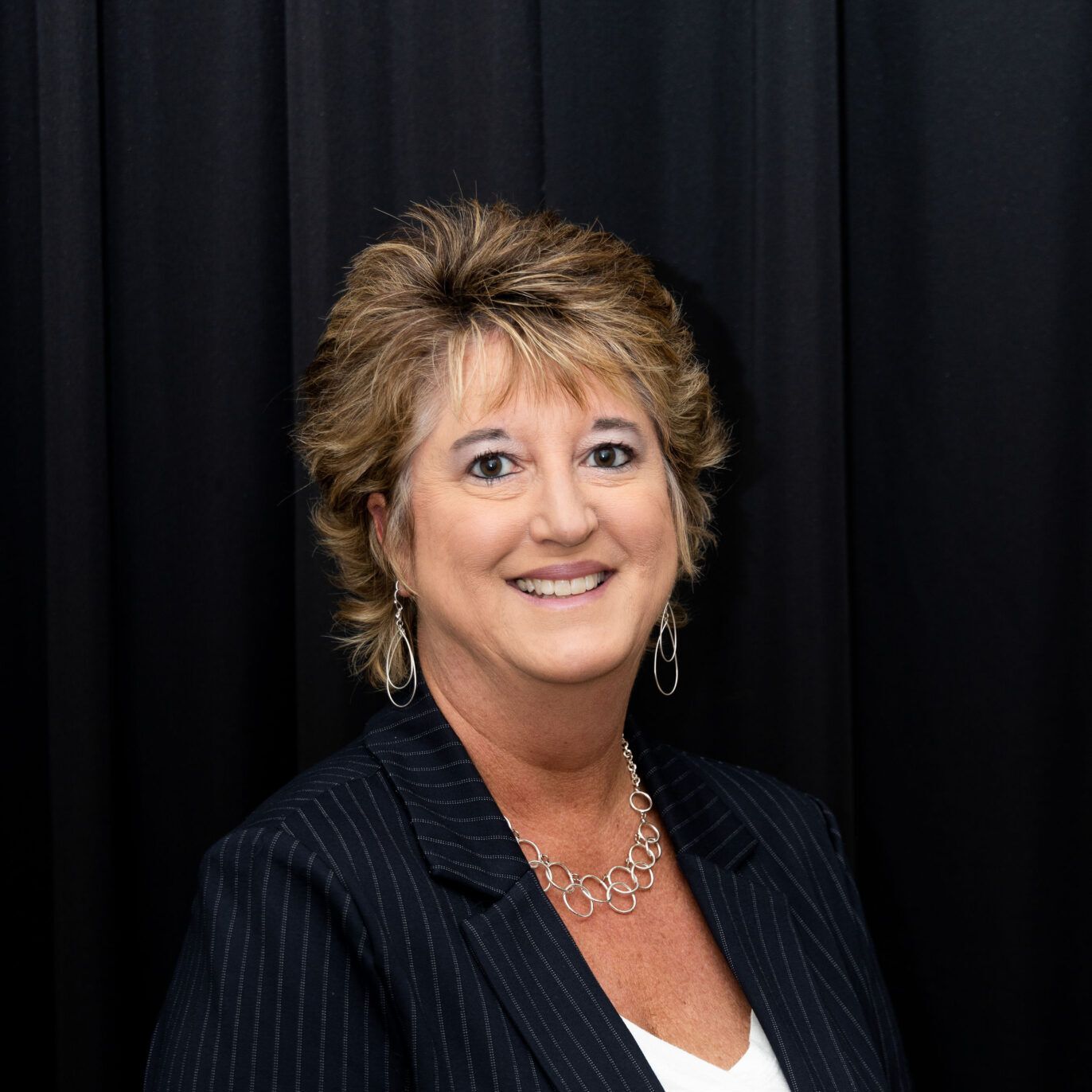 Woman with reddish-brown hair, wearing a black blazer and necklace, smiling against a gray backdrop.