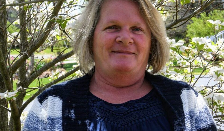 Woman wearing glasses and blazer, smiling. Against a blue background.