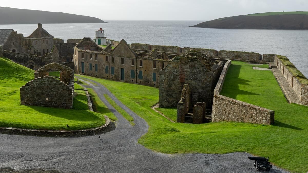 Stone ruins and a grassy courtyard overlooking the sea, with a narrow path and distant shoreline.