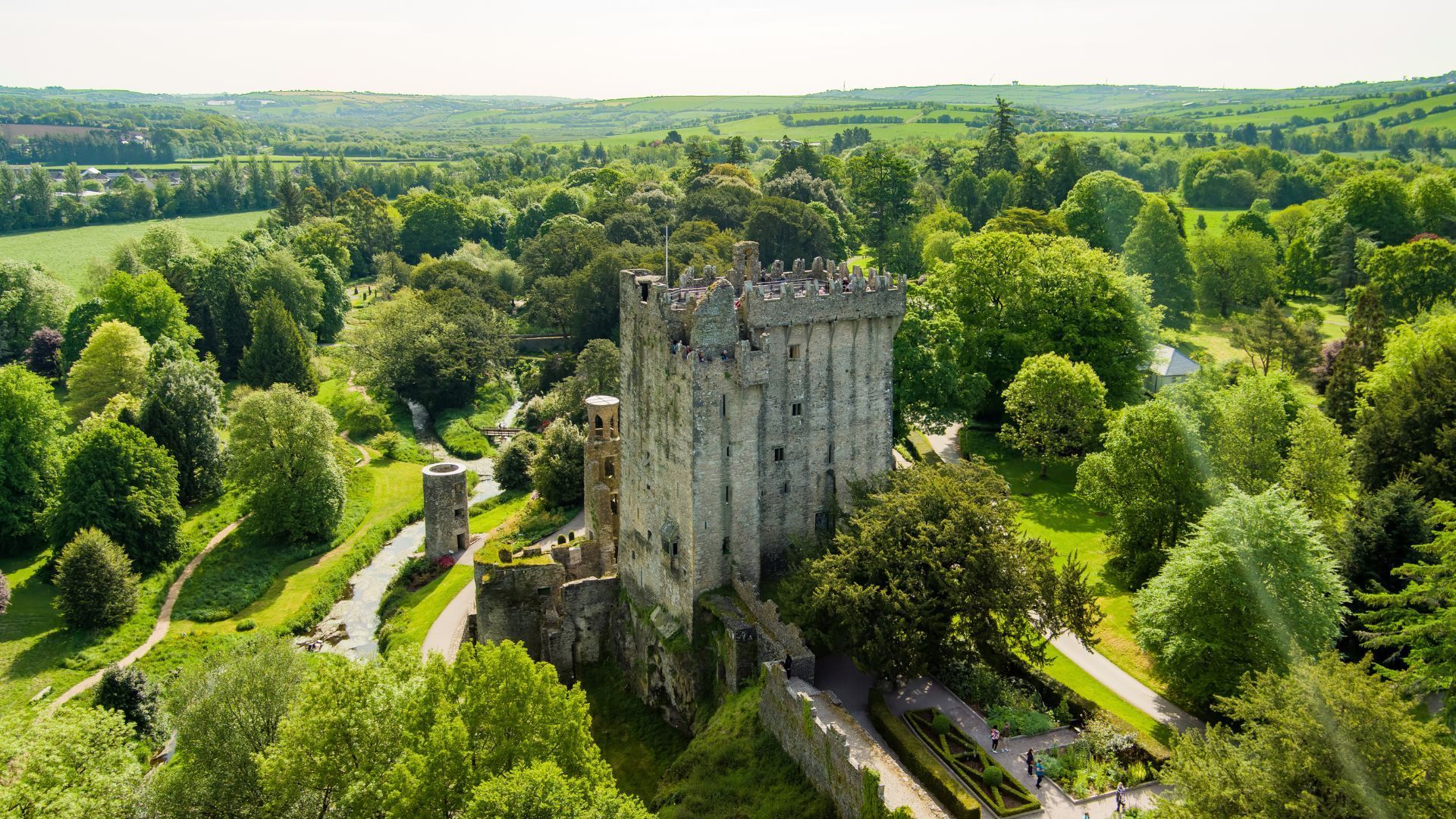 Blarney Castle in Ireland surrounded by lush green trees and gardens under a bright sunny sky.