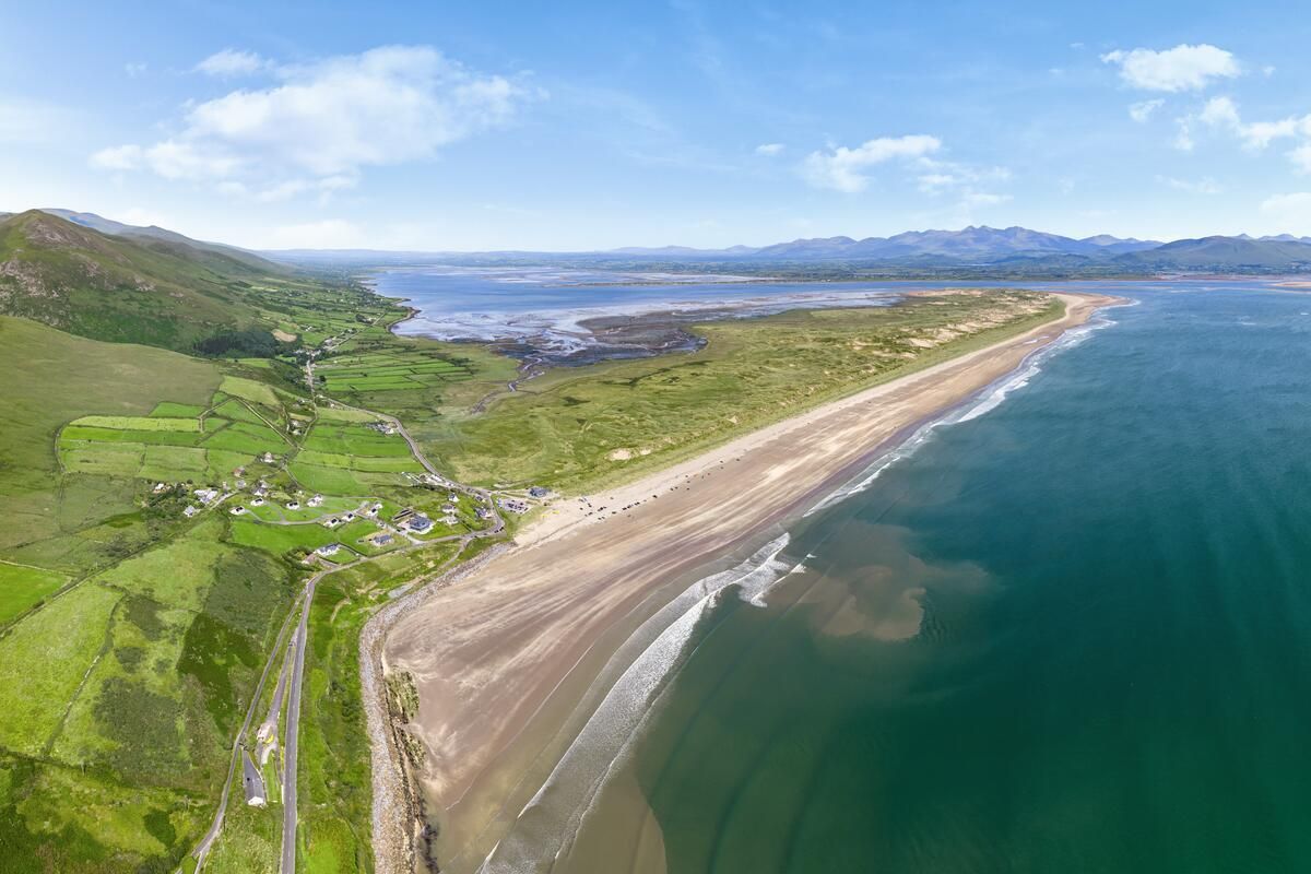 Aerial view of a long sandy beach beside blue water and green hills under a clear sky