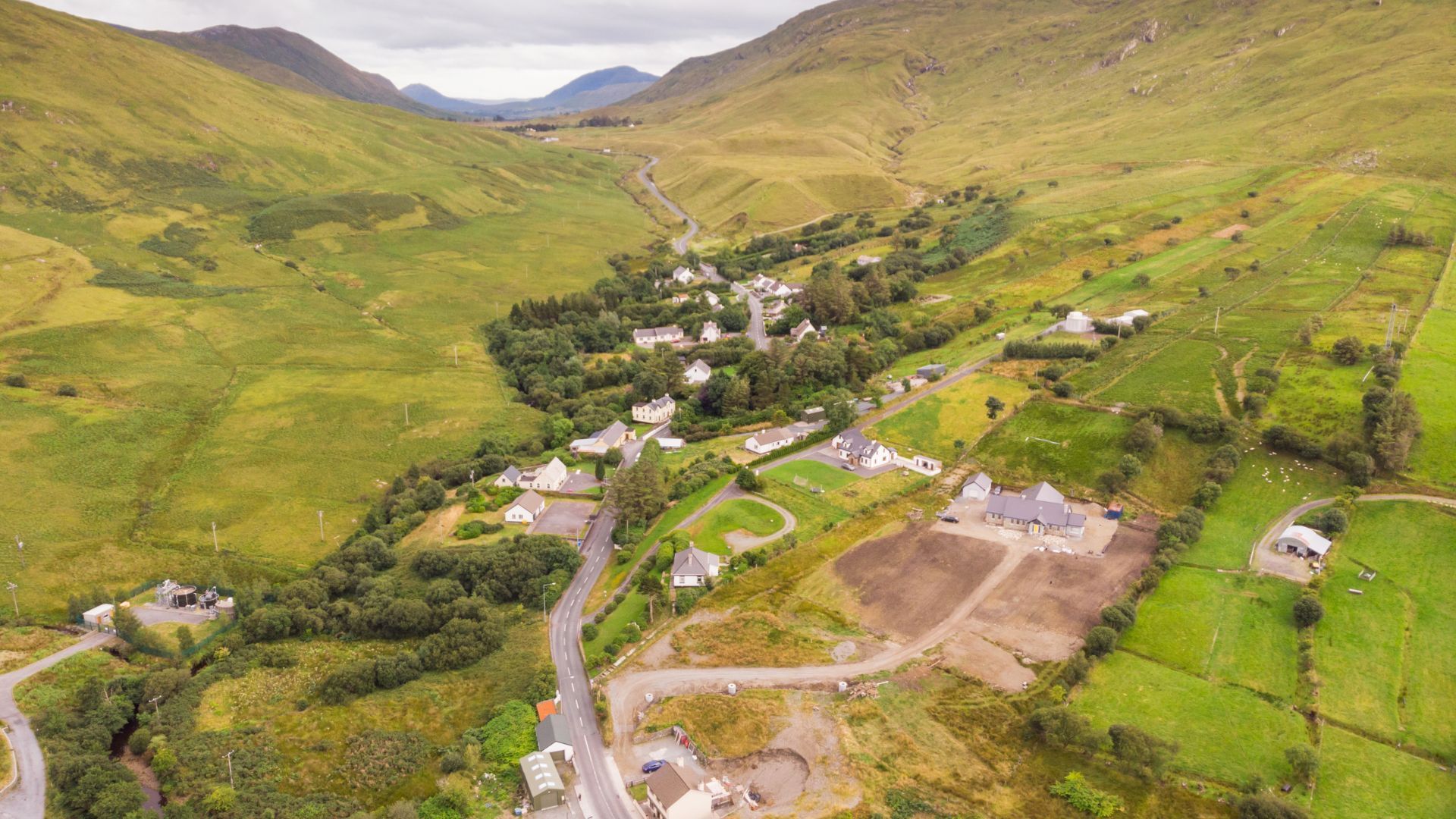 Aerial view of a village nestled in a green valley, surrounded by hills and a road.