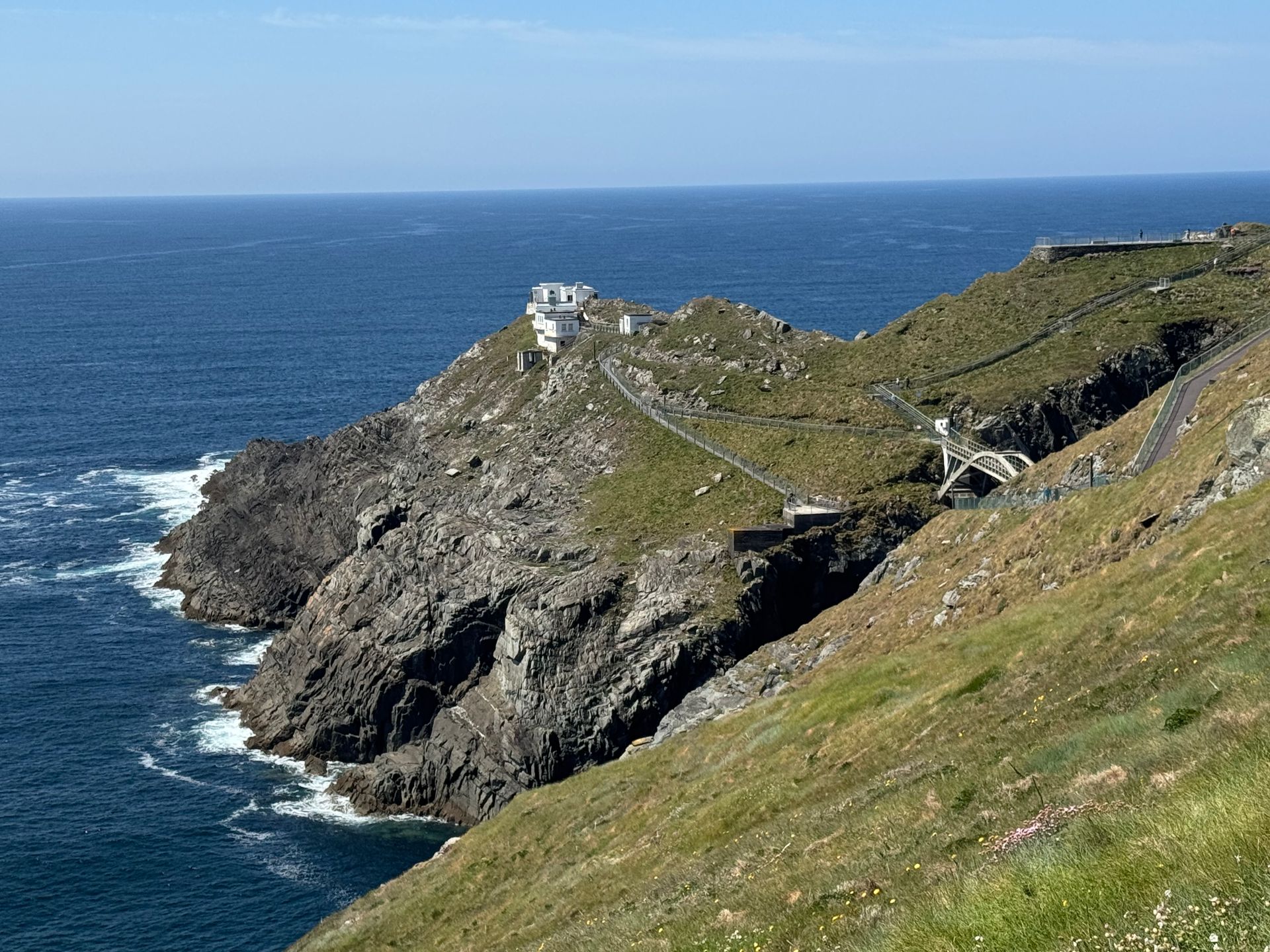 Coastal landscape with a lighthouse atop a rocky cliff, surrounded by ocean, green grass, and blue sky.