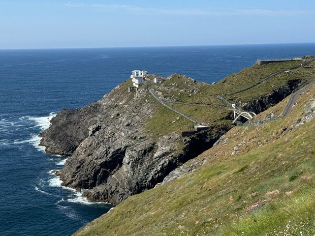 Rocky coastal headland with a lighthouse, pathway, and bridge against a blue sea and sky.