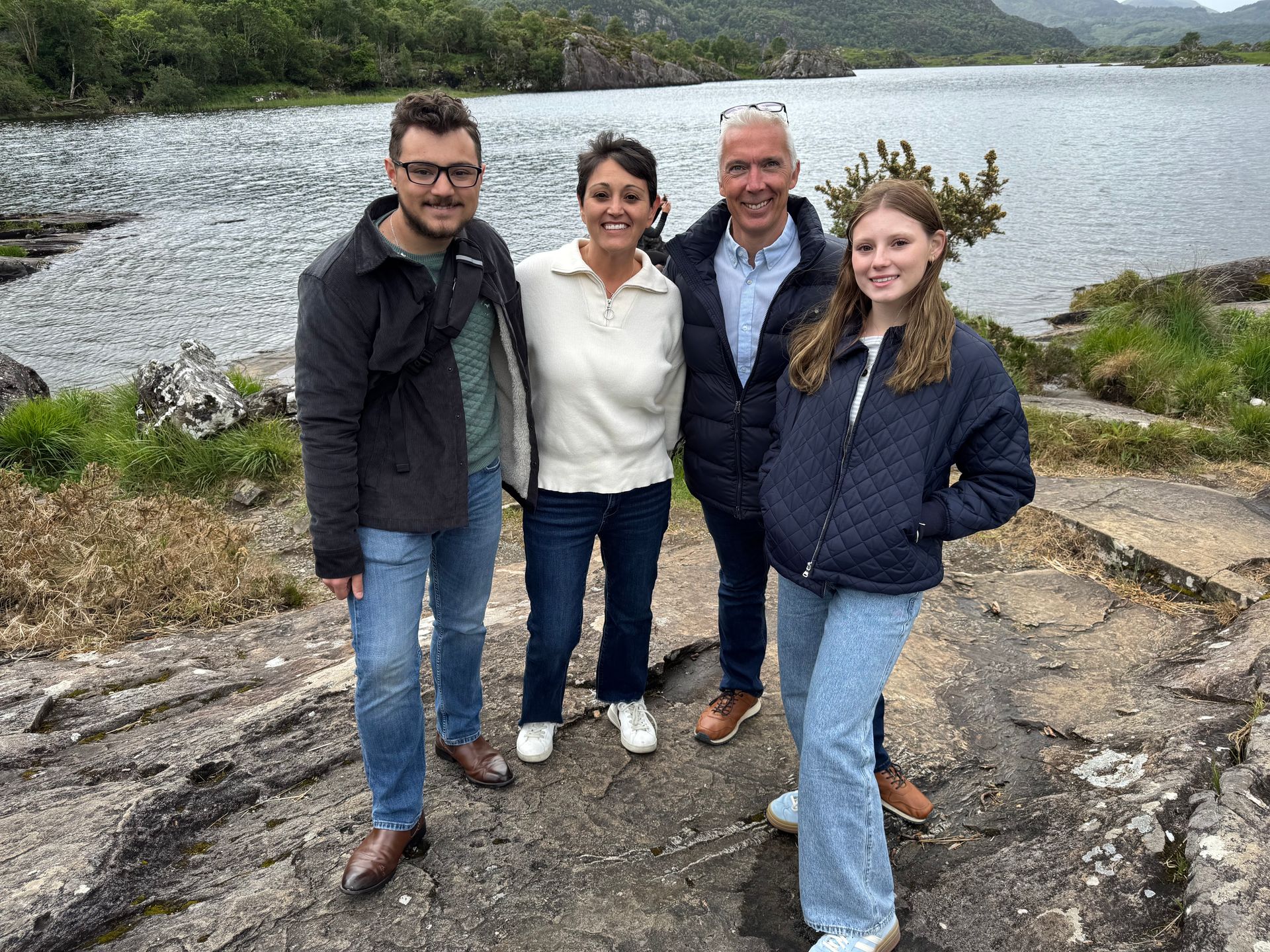 Four people stand on rocks near a lake, smiling.