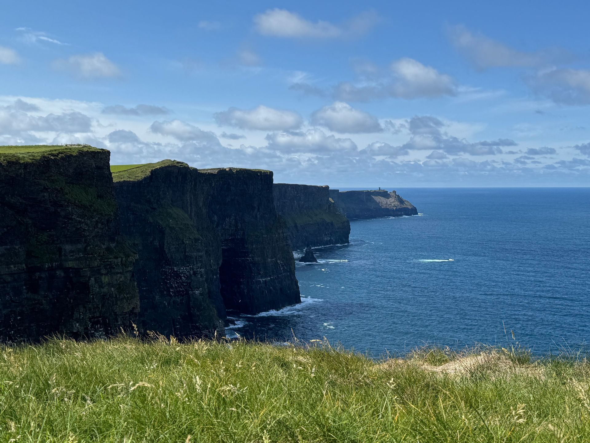 Cliffs of Moher, Ireland, overlooking the blue Atlantic Ocean under a partly cloudy sky. Lush green grass in foreground.