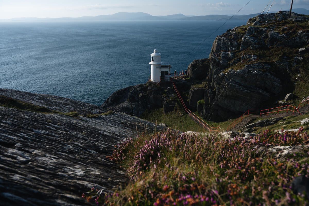 White lighthouse on a rocky cliff above the blue ocean under a hazy sky.