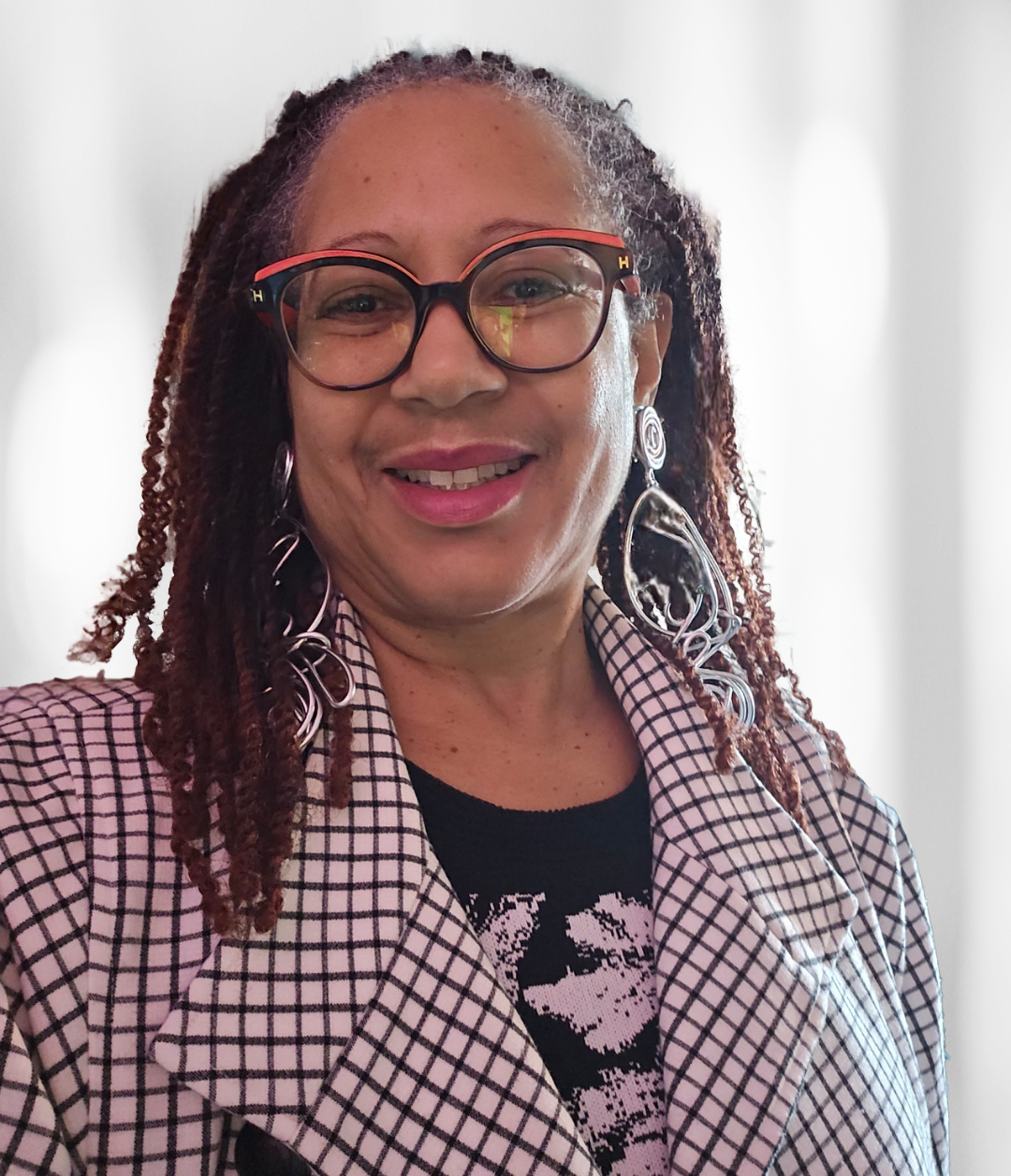 Woman with dreadlocks wearing glasses, a black top, a black and white blazer, and large earrings smiles at the camera.