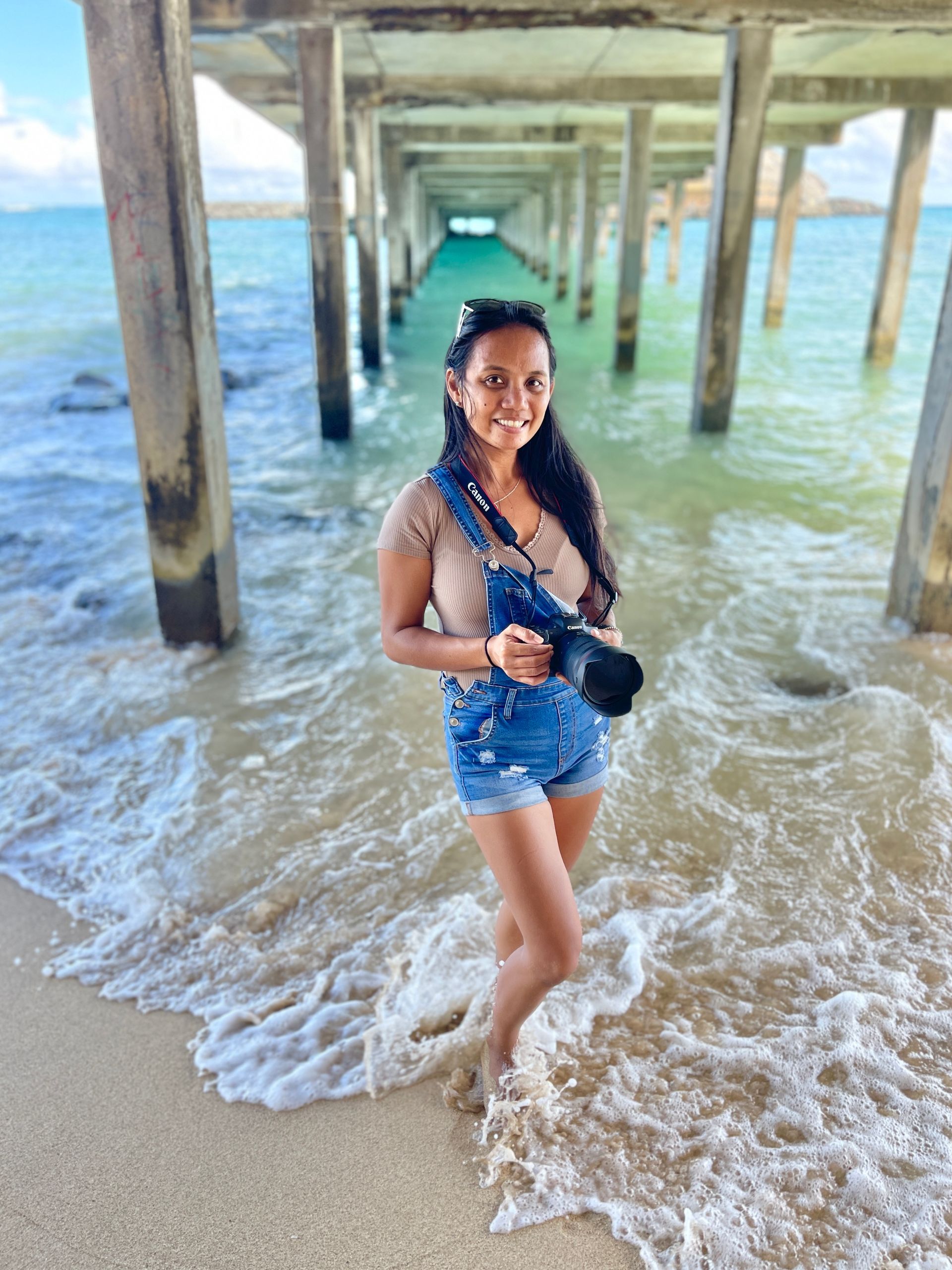 Woman with camera stands in surf under pier. Turquoise water, blue overalls, tan top, smiling.