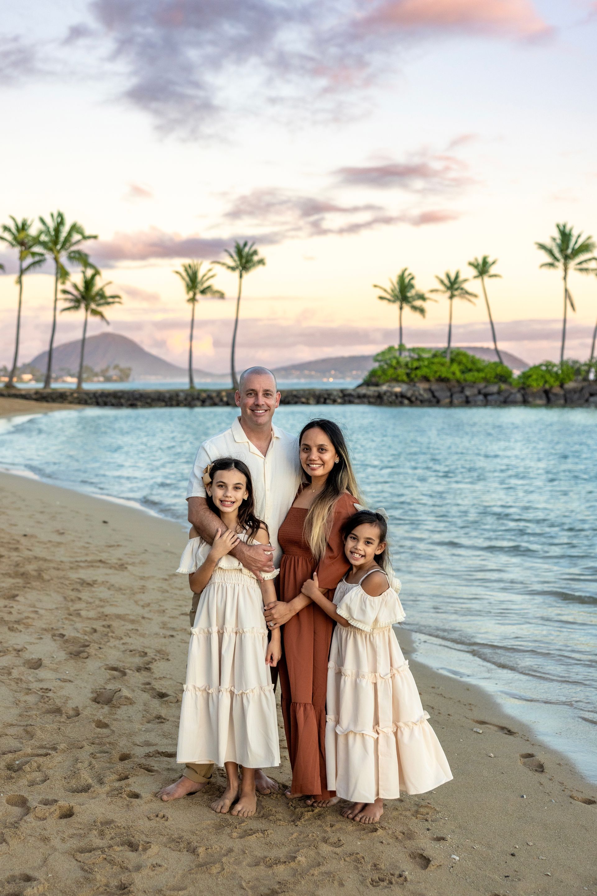 Family of four smiles on a beach at sunset with palms and ocean.