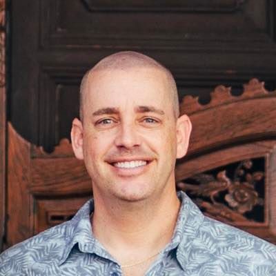 Smiling man with shaved head, wearing a light blue patterned shirt, standing in front of a wooden door.