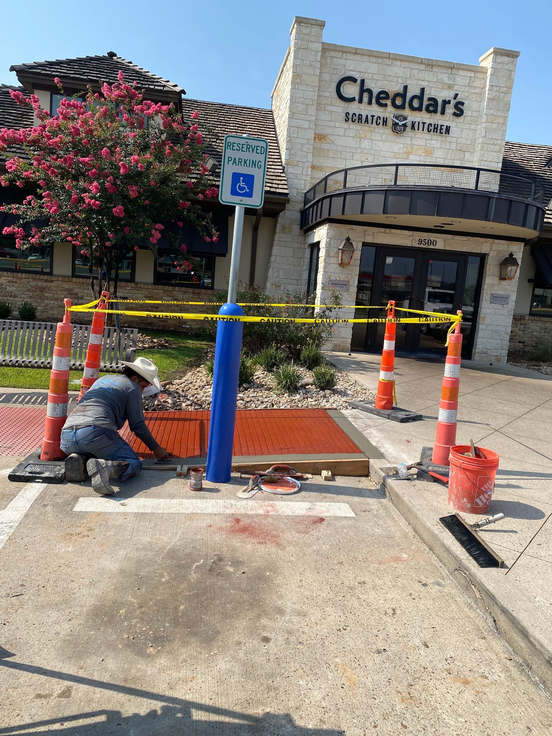 Construction worker repairing a handicapped parking spot at Cheddar's restaurant; orange cones and caution tape present.