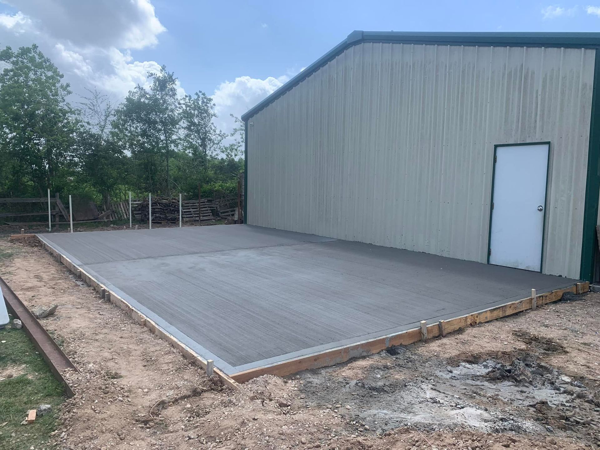 Newly poured concrete slab in front of a metal-sided building. Brown and gray exterior with a white door.