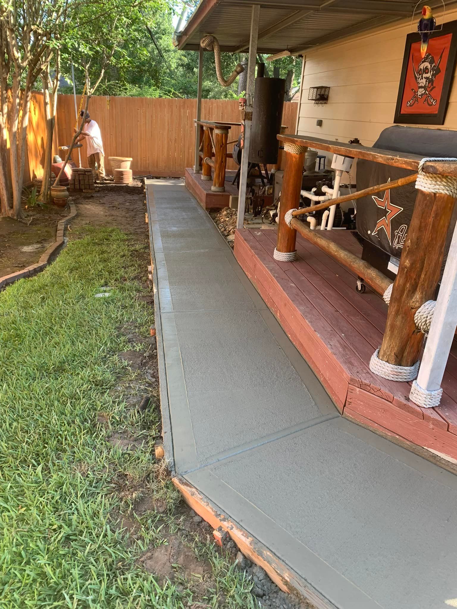Concrete walkway next to a deck and green grass; wooden fence in the background.