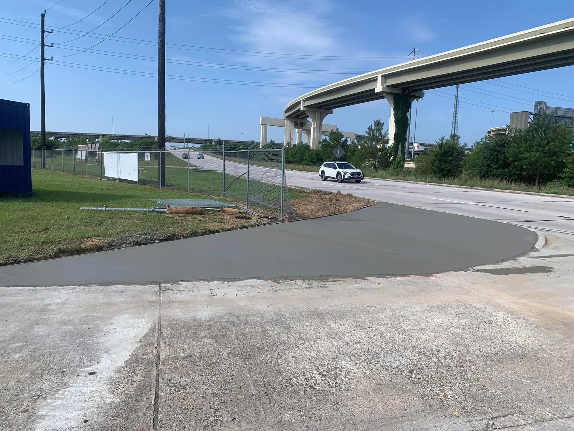 Newly poured concrete on a roadway curve, with a car driving nearby under a bridge.