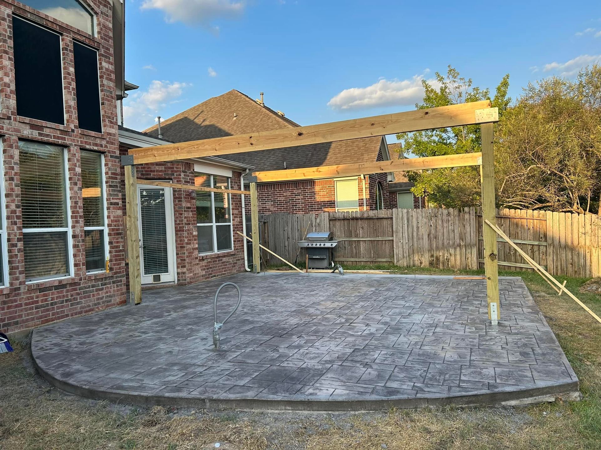 Backyard patio with wooden pergola under construction; stamped concrete patio, brick house, and grill visible.