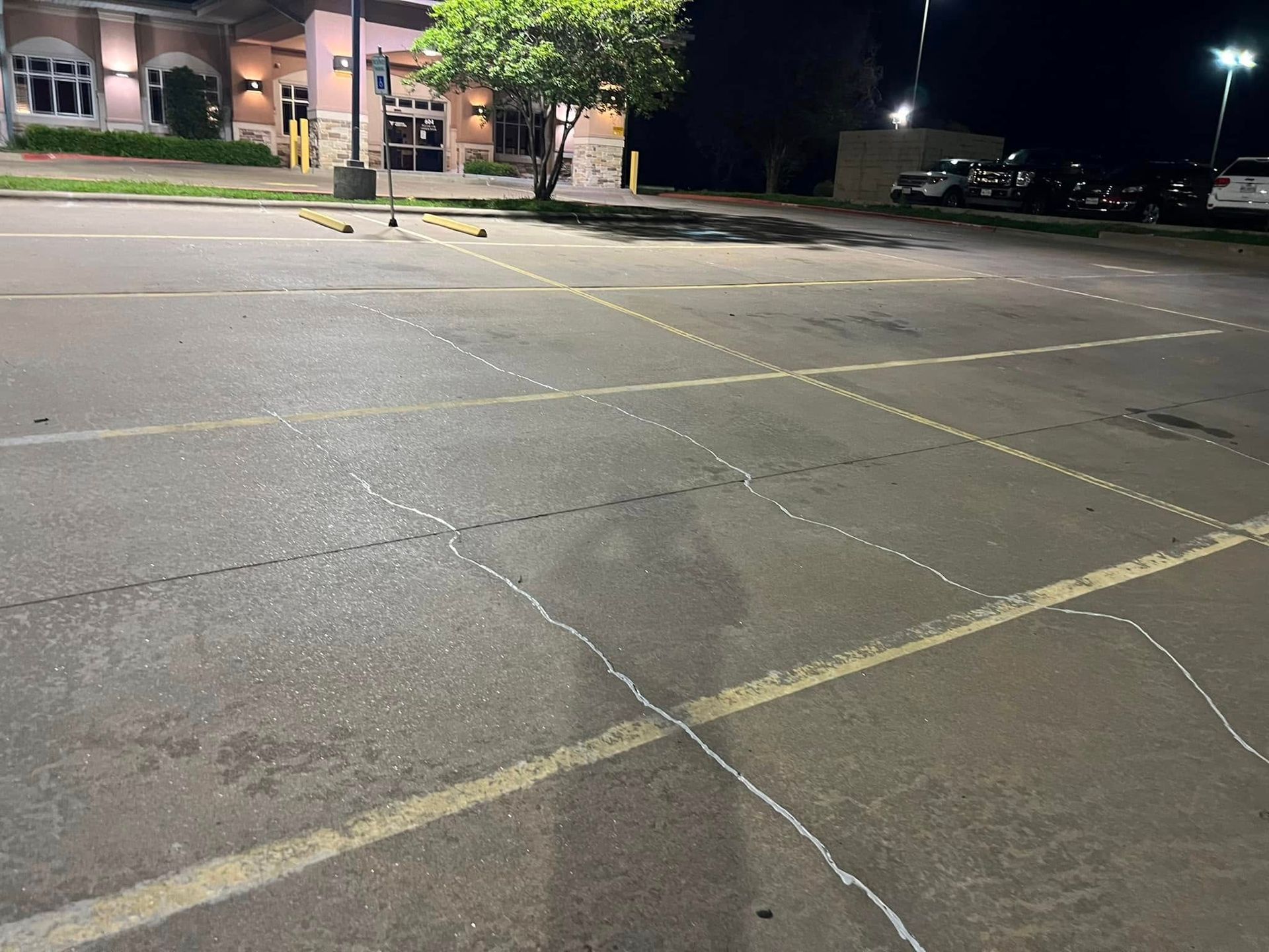 Nighttime view of a cracked asphalt parking lot with painted white lines, a building in the background, and street lights.