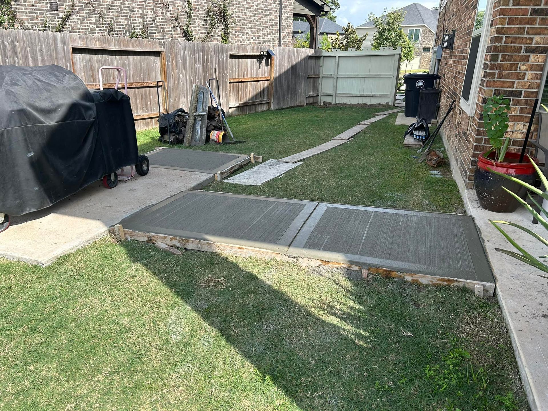 Concrete pathways in a backyard with a grill, fence, and house. Green grass surrounds the walkways.