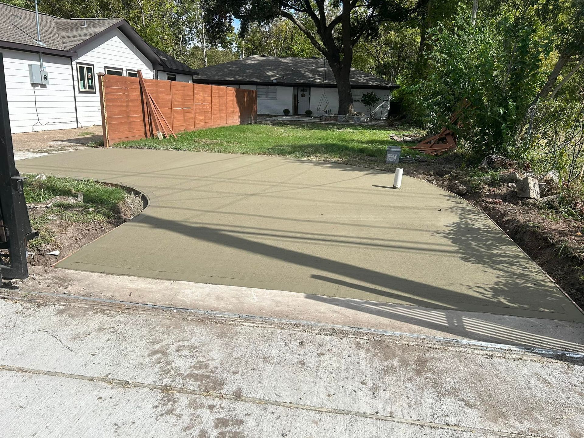 Newly poured concrete driveway extending to a house, surrounded by grass and a wooden fence.