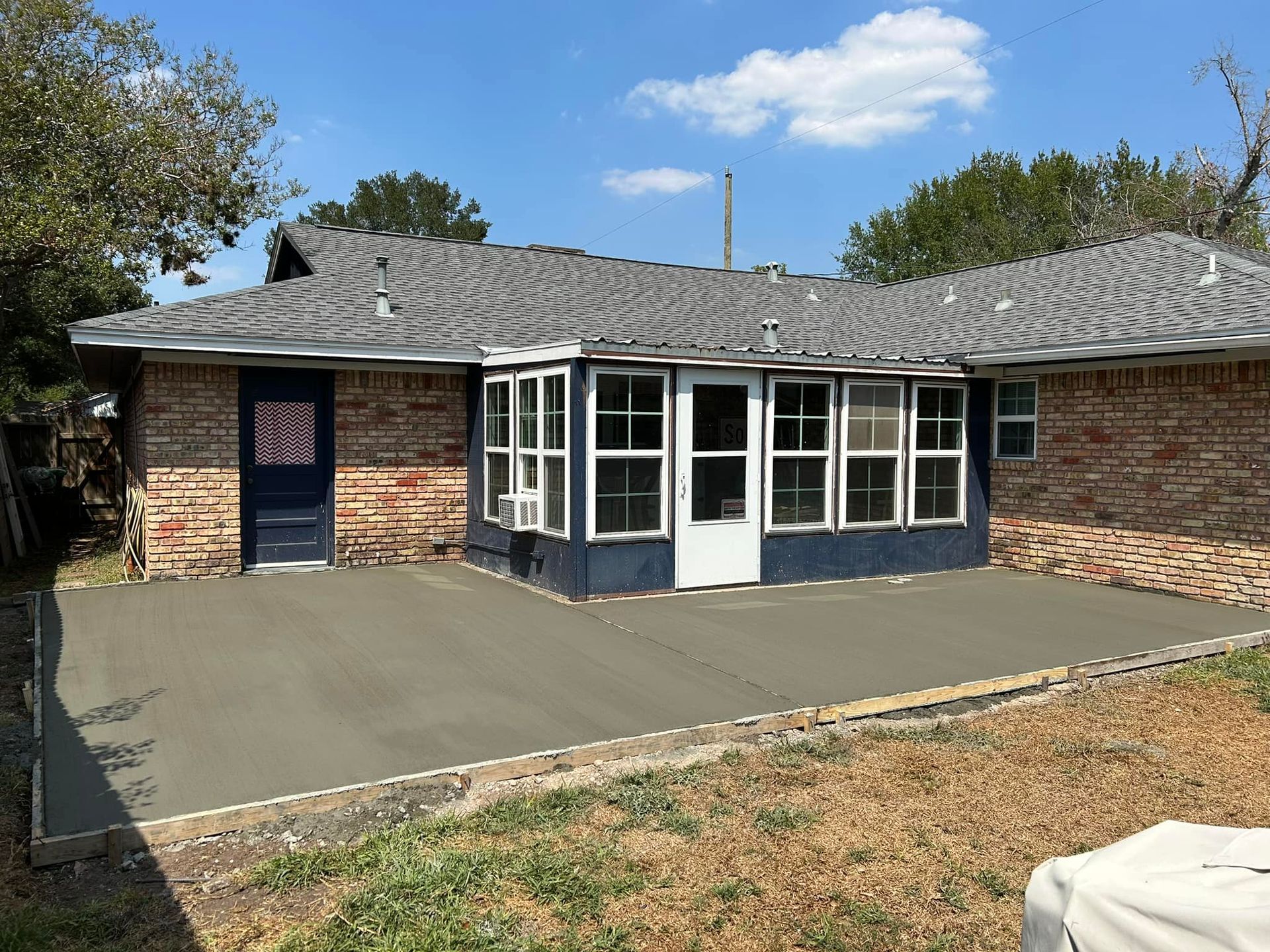 Newly poured concrete patio outside a brick home with an enclosed sunroom and a dark blue door.