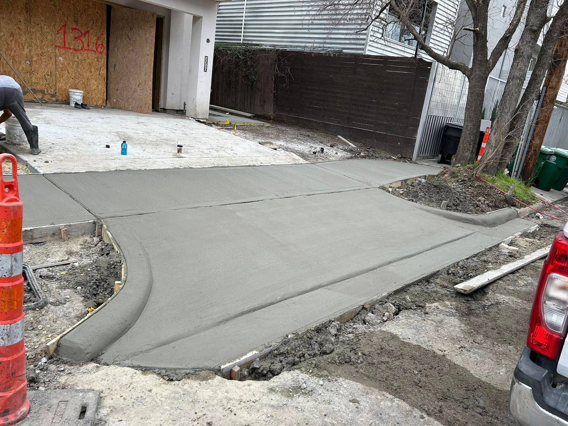 Freshly poured concrete driveway and sidewalk with worker in the background; orange traffic cone in the foreground.