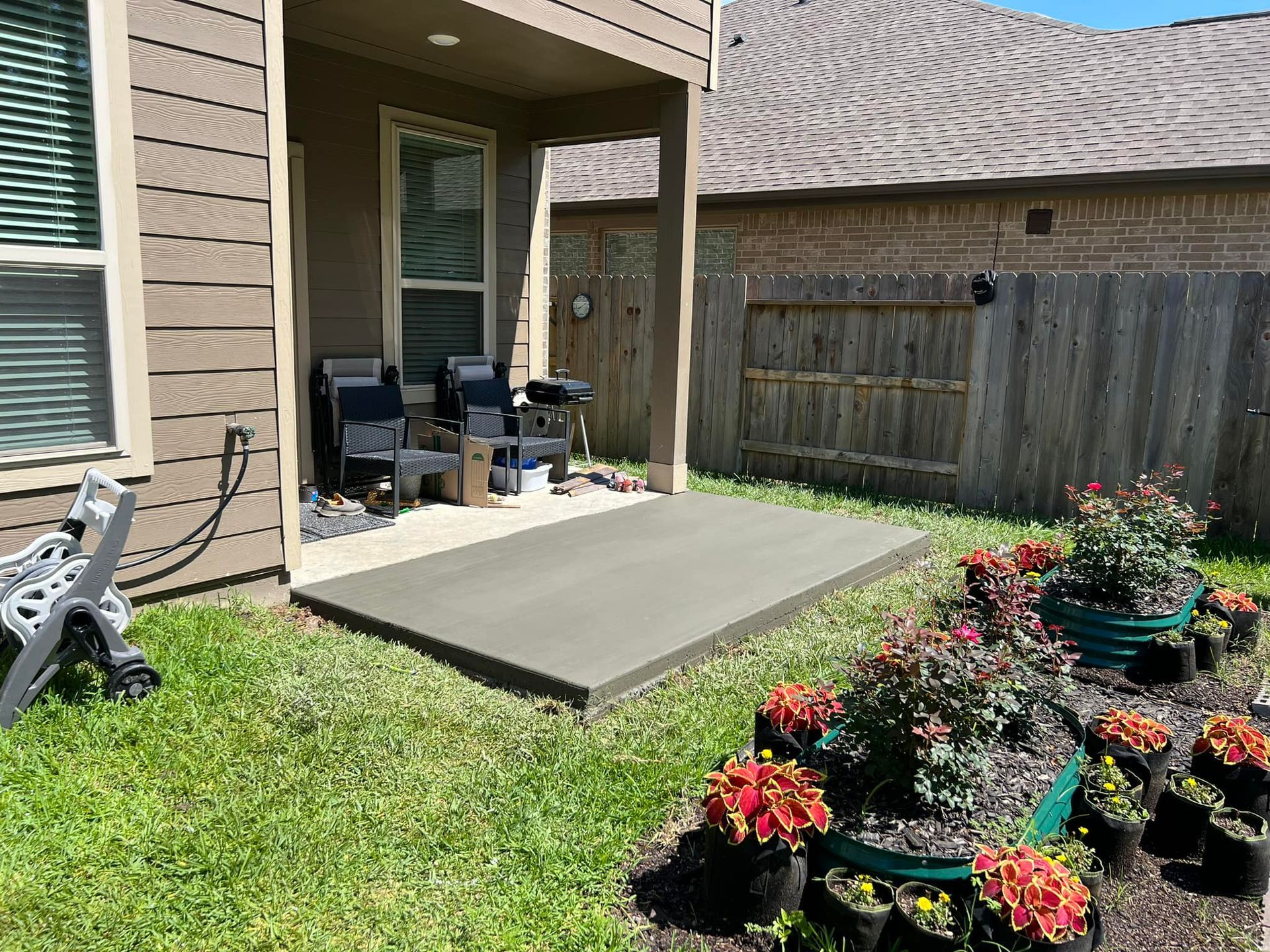 Concrete patio adjacent to house, with grill and chairs. Flower pots surround the patio in grass yard.