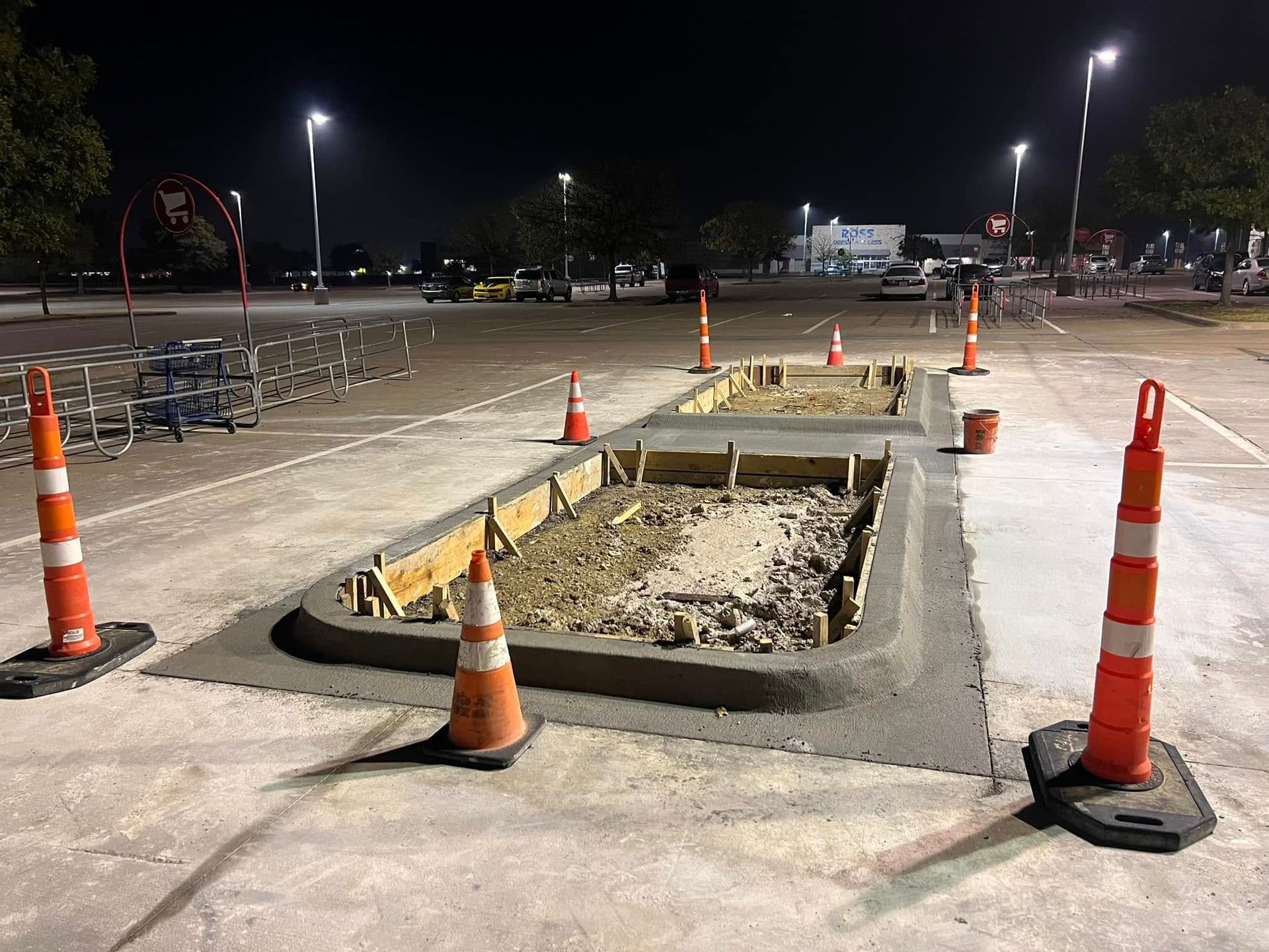 Construction site with concrete forms in a parking lot, marked by orange traffic cones at night.