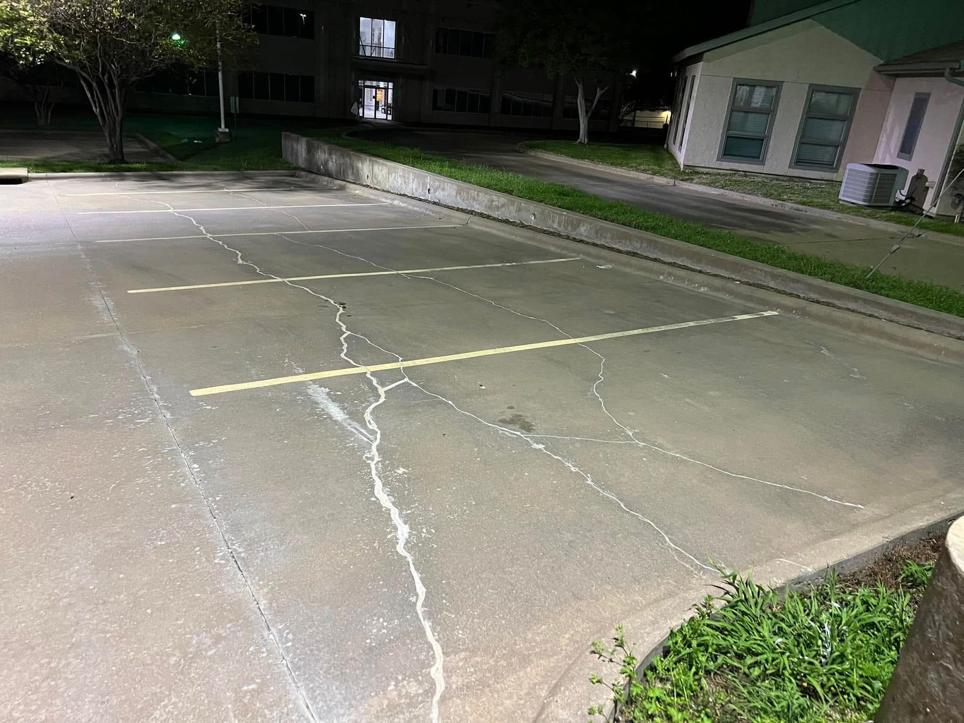 Cracked, empty parking space at night. Yellow lines, building in background, green grass at the bottom.