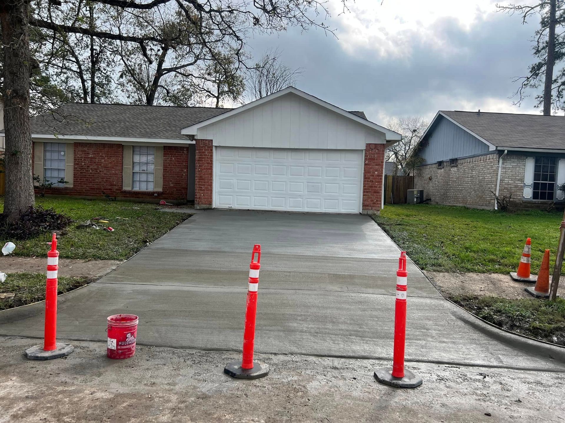 Newly poured concrete driveway in front of a brick house. Red traffic cones mark the perimeter.