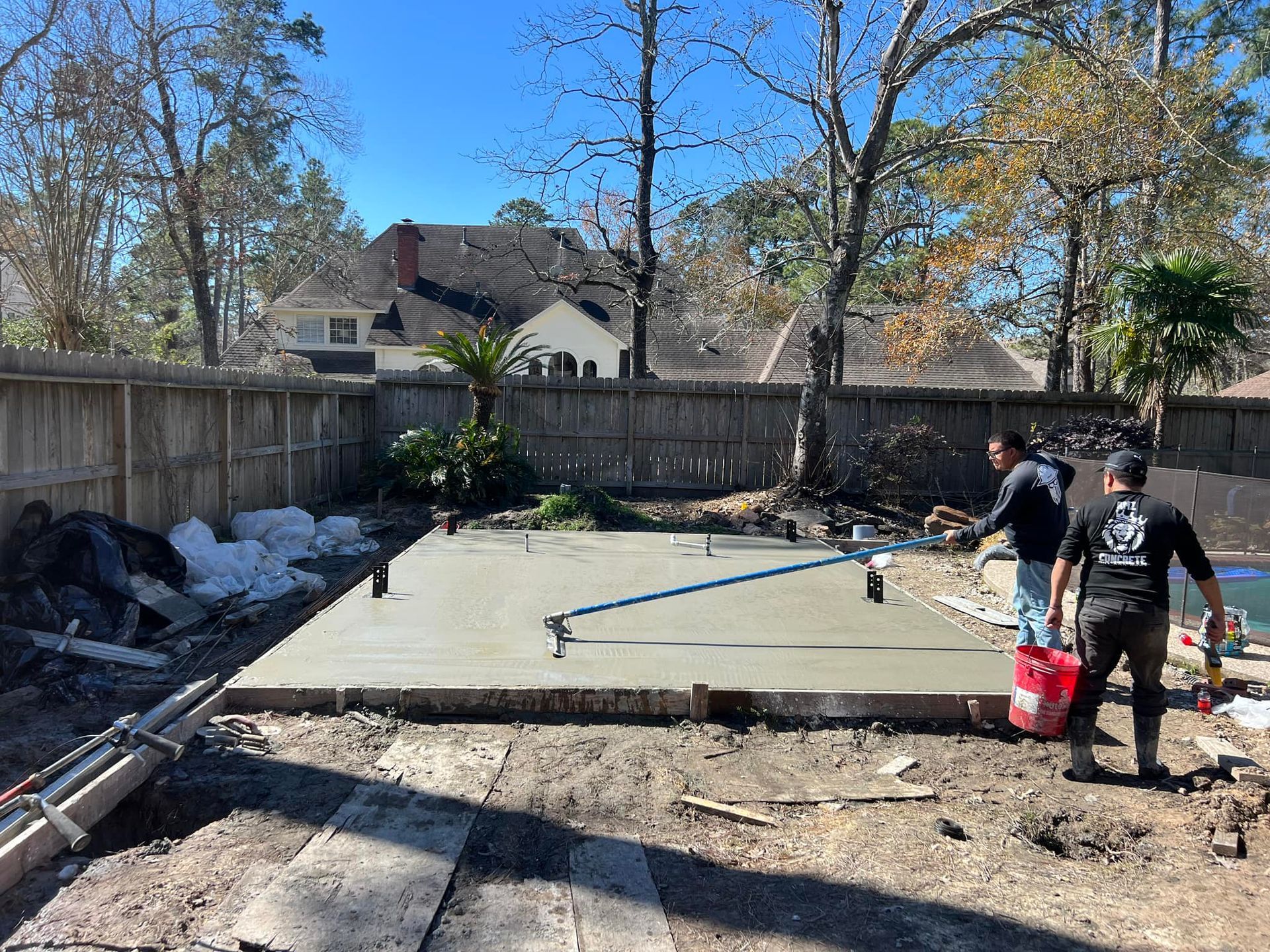 Two workers finishing a concrete slab in a backyard near a fence.