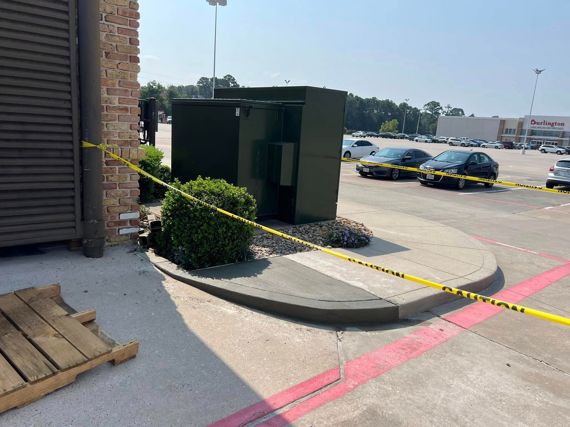 Yellow caution tape surrounds a green electrical box, shrubbery, and a curb in a parking lot.
